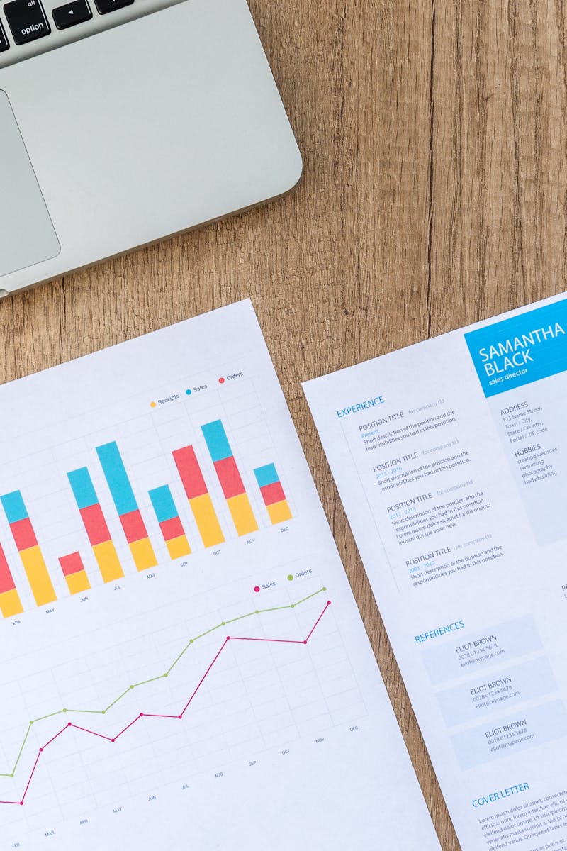 Top view of a laptop, charts, and resume on a wooden desk, showcasing business analysis and job application.